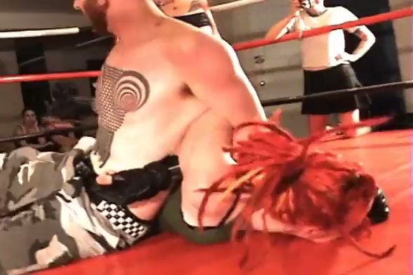 Black-haired dominatrix wields wooden paddle over kneeling opponent in red-walled ring on girlsasian.net